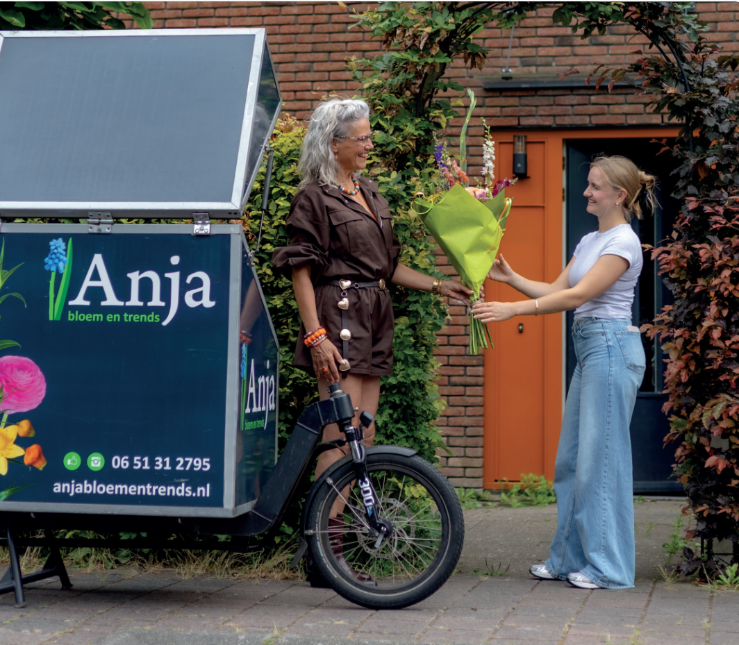 Naamgever Anja van de bloemenwinkel, links in beeld overhandigt een bos bloemen aan een vrouw bij haar huis. Links in beeld een deel van de openstaande elektrische vrachtfiets met daarop de naam en contactgegevens van de winkel.