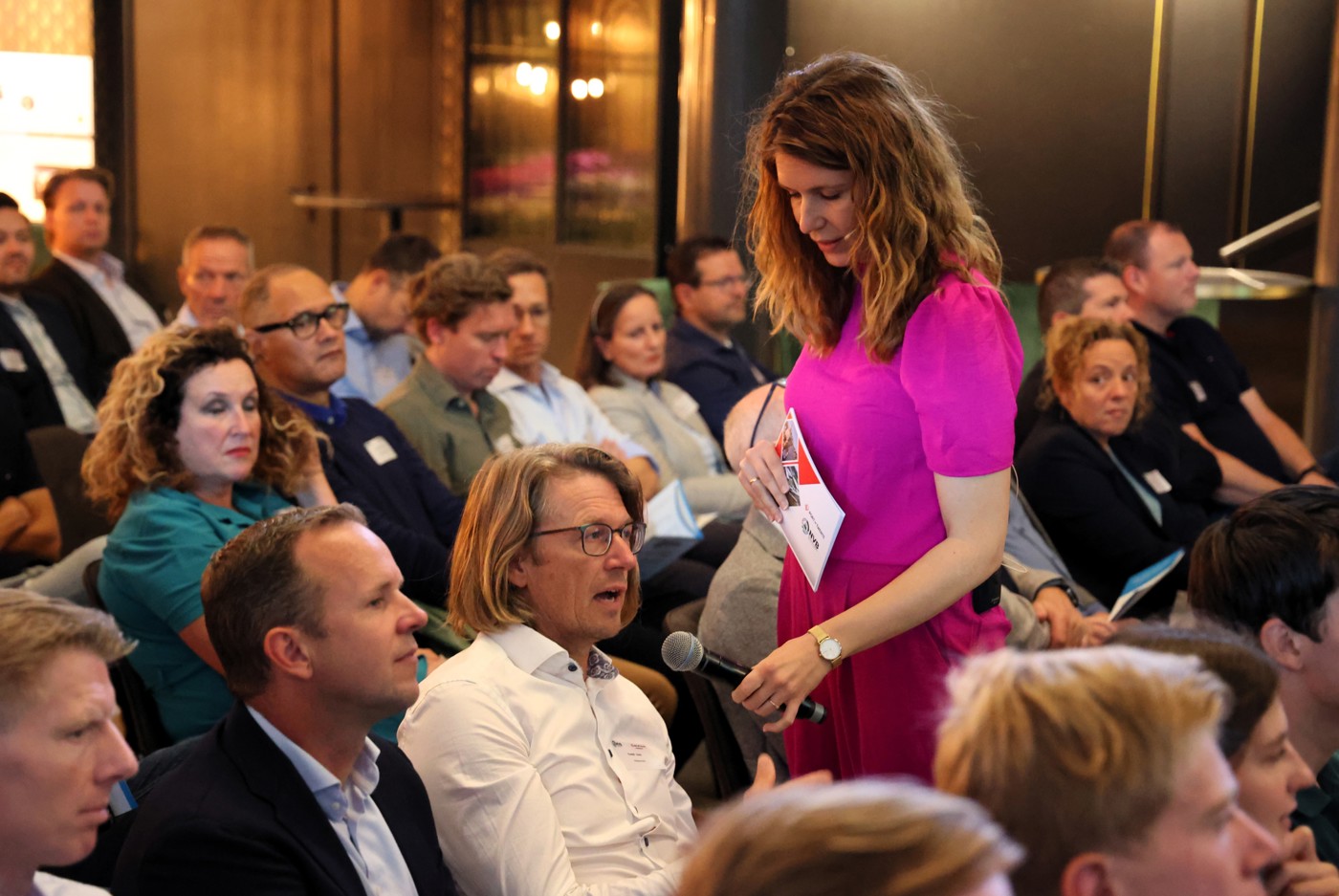 De dagvoorzitster in een opvallend roze pak laat mensen in de zaal aan het woord met een microfoon. 