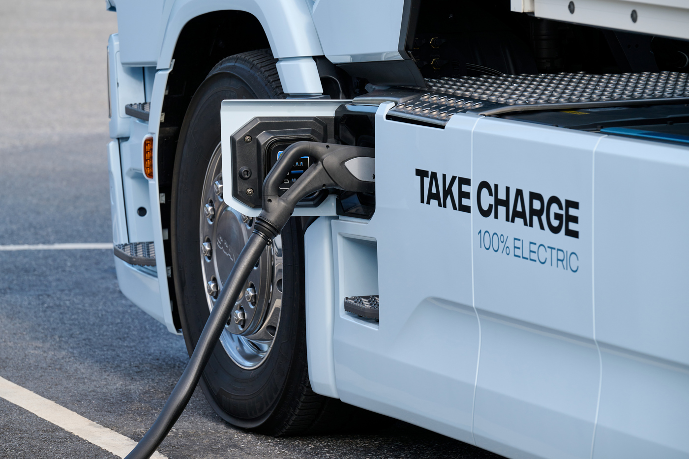 Close-up van witte Scania vrachtwagen die wordt opgeladen. Naast het laadpunt op de vrachtwagen staat de tekst "Take charge, 100% electric".