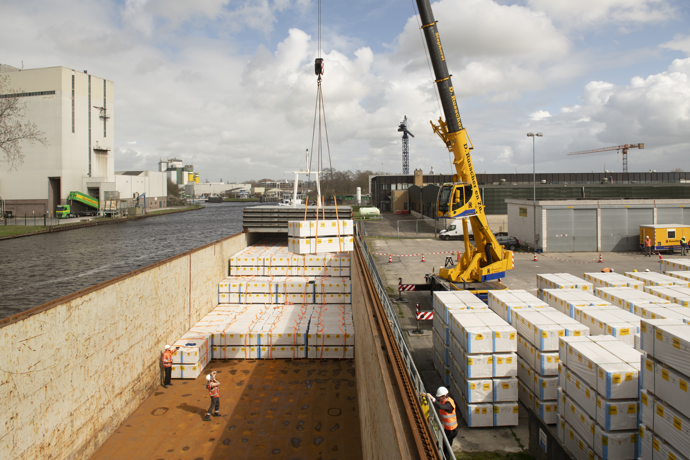 Het kleine zeeschip langs de kade in Meppel wordt beladen met de goederen van Xella door een kraan. In de al half gevulde laadruimte staan ook twee mensen met oranje hesjes en een veiligheidshelm. Een van hen maakt een foto van het beladen van het schip.