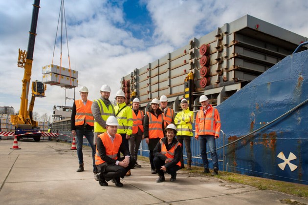 Tien medewerkers en betrokkenen bij Xella poseren met rode en oranje hesjes en witte veiligheidshelmen voor een kruiplijncoaster op de kade bij het bedrijf in Meppel.
